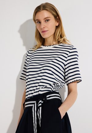 Woman wearing a white and navy striped short-sleeve shirt tucked into black pants with a matching striped belt, one hand in pocket.