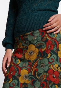 Vihreä neulepusero, jossa on ribatty helma, yhdistettynä moniväriseen kukkakuosiseksi hameeseen, jossa on keltaisia, punaisia ja turkooseja kukkia tummalla taustalla.