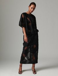 Μαύρο σετ δύο τεμαχίων με διαφανές, υφασμάτινο τοπ με κυματοειδείς υφές και μακριά φούστα με κοψίματα. Πέδιλα με λουράκια.