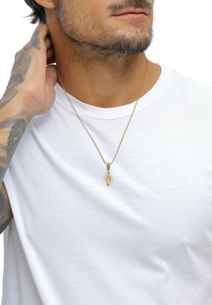 Homme avec une barbe portant une chemise blanche et un collier avec un pendentif cactus en or, touchant son cou avec sa main gauche tatouée.