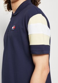 Navy modrá polo košile s texturovaným pletením, která má krátké rukávy s lehce žlutým vzorem a bílým pruhovým akcentem. Logo umístěné na hrudi.