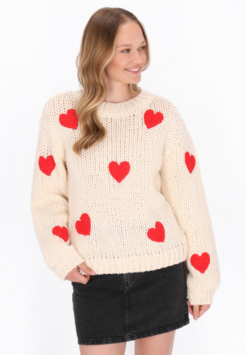 Cremfarbener gestrickter Pullover mit roten Filzherzen-Applikationen, Rundhals und lockerer Passform; kombiniert mit einem schwarzen Jeansrock. Sichtbare Texturdetails im Garn.