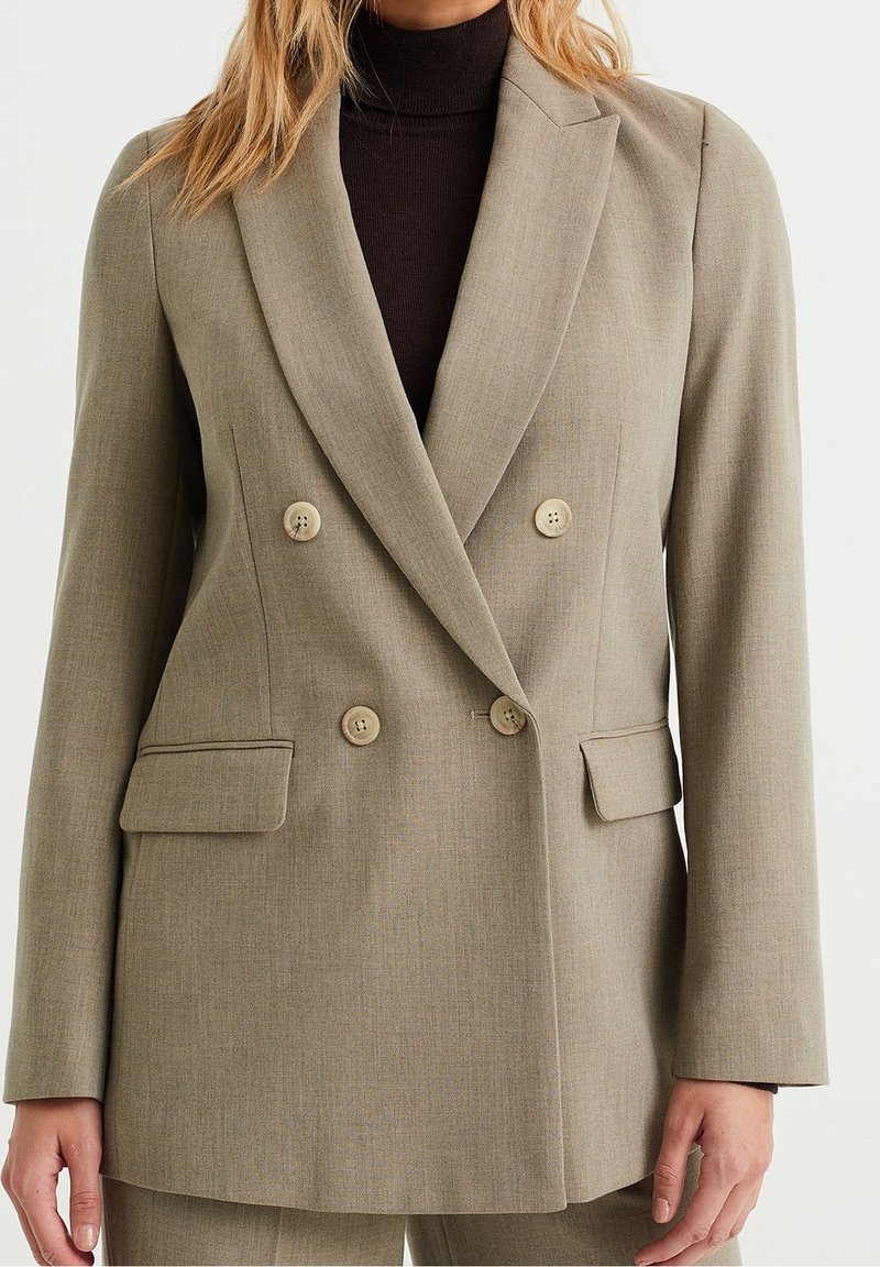 Beige dubbelknäppt blazer med skräddarsydd passform, två framfickor och ljusa knappar. Tillverkad av strukturerad tyg som ger en väldefinierad look.