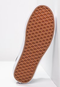 Semelle d'une basket blanche avec un motif gaufré en caoutchouc marron appuyée contre un mur d'angle blanc sur une surface blanche.