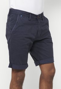 Man in marineblauwe opgerolde shorts en een wit shirt, afgebeeld van taille tot halverwege de dij op een effen achtergrond.