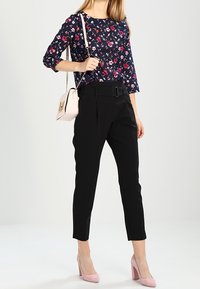 Kobieta nosząca czarne, wysokie spodnie z paskiem, granatową kwiecistą bluzkę, różowe szpilki i jasnoróżową torebkę na ramię z złotym łańcuszkiem.