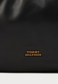 Czarna skórzana torebka z fakturowanym wykończeniem i wytłoczonym złotym logo z napisem "TOMMY HILFIGER" na dole.