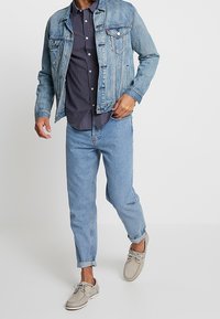 Giacca di jeans di un blu chiaro, camicia a fantasia sotto, jeans slim fit di un blu chiaro e mocassini grigi con lacci e punta tonda.