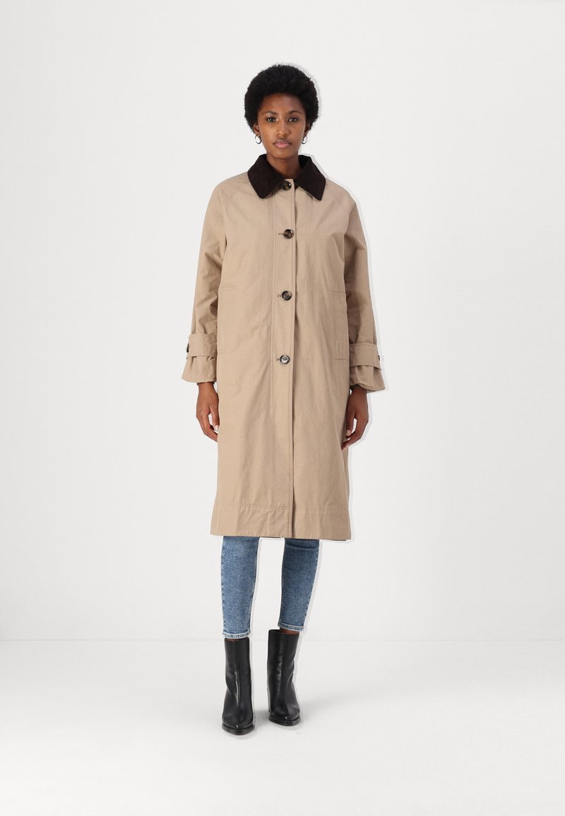 Beige trench coat barna gallérral, elülső gombokkal és mandzsetta részletekkel. Kék farmernadrág és fekete bokacsizma fölé viseled; sima textúra.