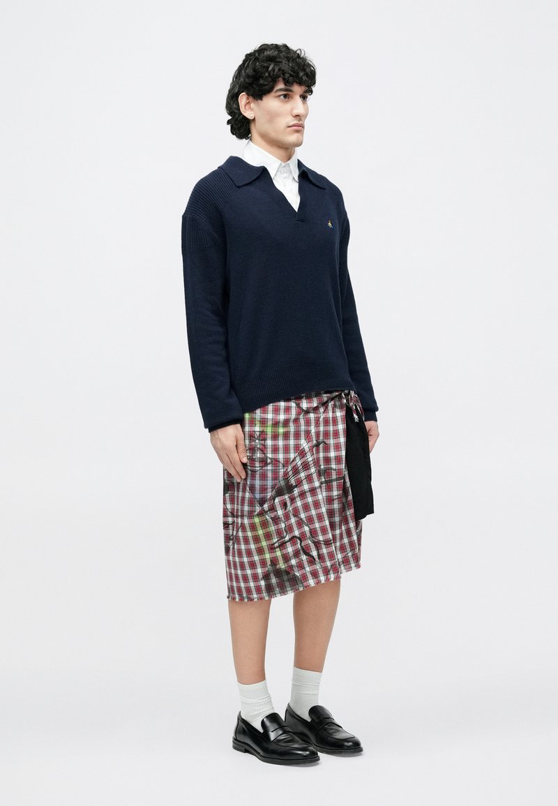 Un homme porte un pull bleu marine sur une chemise blanche, une jupe plissée rouge à carreaux longueur genou avec des détails noirs et jaunes, des chaussettes blanches et des mocassins noirs.