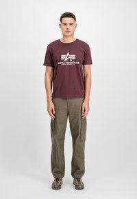 Burgunder T-Shirt mit weißem "Alpha Industries" Logo, kombiniert mit olivfarbenen Cargo-Hosen und Schuhen im Tarnmuster. Einfaches Rundhals-Design.