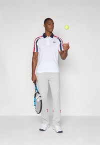 Weißes Poloshirt mit marineblauem Kragen, roten und marineblauen Streifen an den Ärmeln; graue Sporthose; hält einen Tennisschläger und jongliert mit einem Tennisball.