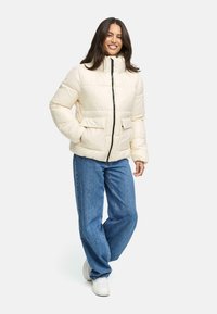 Puffjakke i creme, med høj krave, frontlynlås og to sidelommer. Bæres med afslappede blå jeans og hvide sneakers.