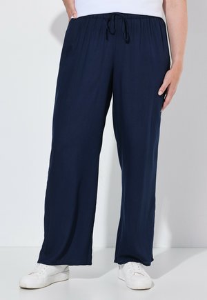 Person trägt dunkelblaue weit geschnittene Hose mit elastischem Bund und weiße Sneaker, steht vor einem einfarbigen hellen Hintergrund.