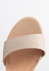 Beige sandaali leveän kimaltavan kangasnauhan ja sileän, pyöreän varpaan kanssa. Pohja on vaalean värinen ja hieman teksturoitu.