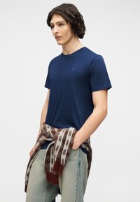 Νέος άνδρας που φοράει μπλε μπλουζάκι με μικρό λογότυπο, ανοιχτόχρωμο τζιν και καφέ ριγέ πουλόβερ δεμένο γύρω από τη μέση του.