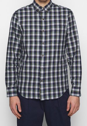 Hombre con camisa de manga larga abotonada con patrón de cuadros en azul marino, blanco y verde, combinada con pantalones azul marino oscuro.
