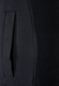 I pantaloni tuta neri in tessuto presentano una tasca laterale con un'apertura arrotondata, una texture liscia e dettagli di cucitura visibili lungo le cuciture.