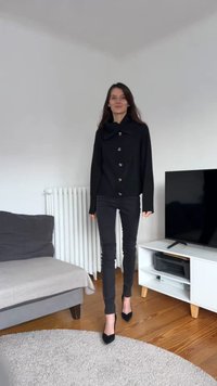 Melnā pogājama jaka ar lielu apkakli, kombinēta ar melnām šaurām biksēm un melniem smailiem papēžiem, iestatīta minimālisma interjerā.