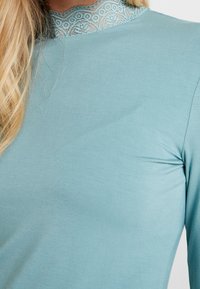Light teal long-sleeve top with a textured lace neckline, featuring soft fabric and a fitted design, creating a smooth silhouette.