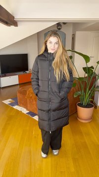 Hosszú fekete pufi kabát cipzárral elöl, kapucnival és oldalzsebbel, fekete nadrágra és ezüst sportcipőre viselve. Beltéri környezet növényekkel.