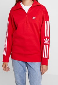 Röd fleece-pullover med kvartdragkedja, vitt Adidas-logotyp på bröstet och tre vita ränder på varje ärm, ihop med blå jeans.