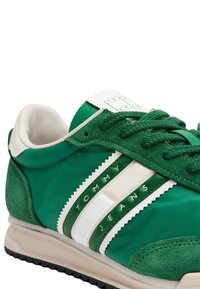 Zapatillas verdes con una mezcla de ante y material sintético, con rayas blancas y el texto "TOMMY JEANS". Suela de goma con detalles texturizados.