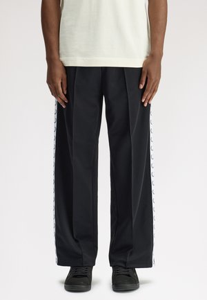 Persona con pantalones negros de pierna ancha con franjas laterales blancas que presentan pequeños logotipos negros, combinados con zapatillas negras y una camiseta de color claro.