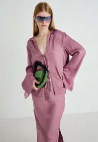 Donna che indossa una blusa e una gonna con rifiniture in pizzo viola, tiene in mano una borsa verde con perline, indossa occhiali da sole blu grandi e orecchini a cerchio.