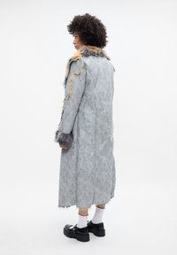 Garšs pelēks mētelis ar kažokādas akcentiem, kuram ir tekstūrēta apdare, plecu detaļas un kažokādas aproces. Apakšā valkā melnus platformas apavus.