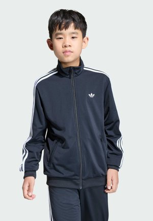 Ragazzo che indossa una giacca sportiva Adidas nera e pantaloni con strisce bianche, in piedi su uno sfondo chiaro e uniforme.