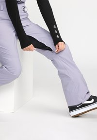Pantalons de neige violet clair avec des aérations zippées. Les caractéristiques incluent une doublure noire texturée et une patch de logo. Associés avec des sneakers noires.