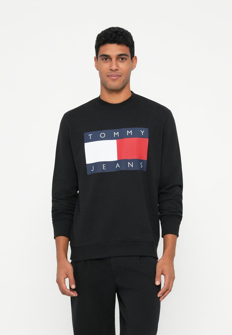 Hombre vestido con una sudadera negra con un gran logotipo de Tommy Jeans en blanco, rojo y azul, de pie contra un fondo blanco liso.