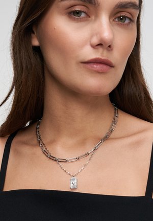 Frau trägt geschichtete silberne Kettenhalsketten, eine mit länglichen Gliedern und die andere mit einem rechteckigen Anhänger, gekleidet in einem schwarzen Oberteil.