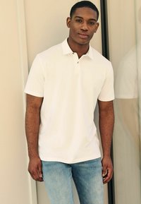 Camisa polo blanca con cuello, mangas cortas y tapeta de tres botones, combinada con unos pantalones vaqueros de denim claros. Tejido de algodón suave.