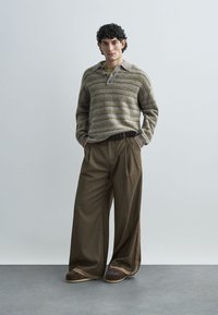 Gestreifter Strickpullover in Grün und Beige, kombiniert mit weiten braunen Hosen mit seitlichem Streifendetail. Braune Schuhe vervollständigen das Outfit.