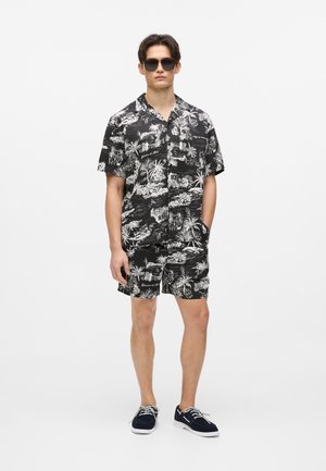Zestaw koszuli na krótki rękaw i szortów z tropikalnym, czarno-białym nadrukiem, wykonany z lekkiej tkaniny, z przednimi kieszeniami i swobodnym krojem. Założone okulary przeciwsłoneczne.