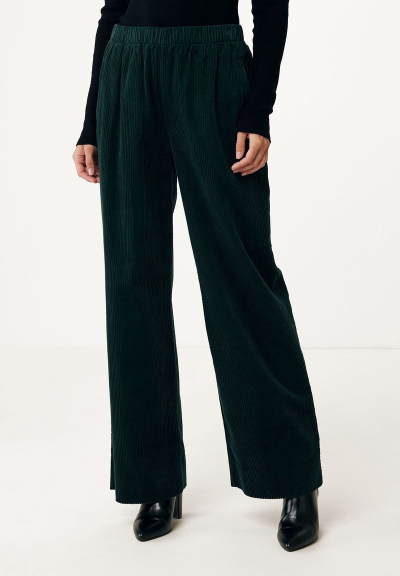 Pantalon large en velours côtelé vert foncé avec taille élastique porté avec des bottines noires à talons et un haut noir à manches longues.