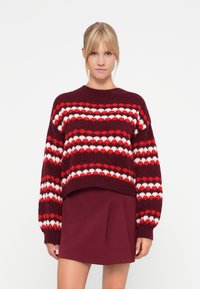 Knitted sweater in burgundy with red and white heart patterns, thick ribbed cuffs, and a cropped design. Paired with a burgundy pleated skirt.