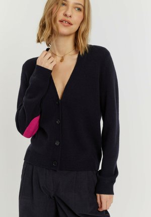 Vrouw die een zwart vest met knopen en een roze ellebooglap draagt, en een zwarte broek, terwijl ze een gouden hangerketting aanraakt.
