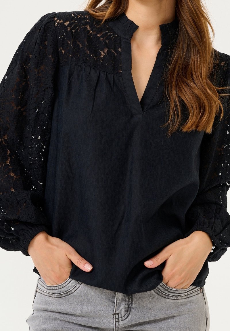 Blouse noire avec manches en dentelle et col mandarin. Tissu légèrement texturé, coupe ample et détails plissés à l'avant.
