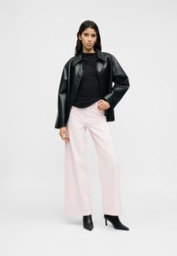 Veste noire en cuir synthétique, haut noir ajusté, pantalon large rose clair, et bottines noires pointues. Le mannequin se tient devant un arrière-plan neutre.