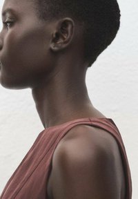 Top sin mangas de color marrón con una tela suave y caída. El cuello y los hombros del modelo se ven lisos, con el cabello corto y muy recortado. Fondo neutro.