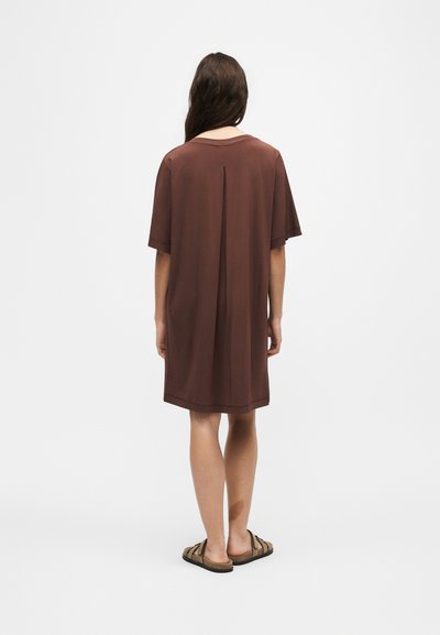 Femme aux longs cheveux bruns portant une robe marron ample jusqu'aux genoux et des sandales marron, debout de dos sur un fond blanc uni.