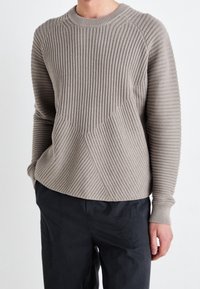 Beiger Rippenpullover mit Rundhalsausschnitt, strukturiertem diagonalen Muster und enganliegenden Manschetten, kombiniert mit dunklen Hosen.