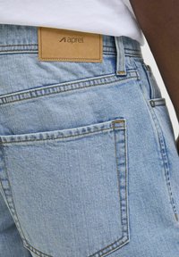 Buzunarul din spate al blugilor din denim albastru deschis, cu cusături vizibile și o etichetă de brand din piele maro pe talie, purtate cu o cămașă albă.