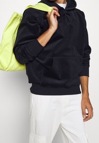 Schwarzer Hoodie mit einer Fronttasche und einer Kapuze mit Kordelzug. Kombiniert mit einer neon-gelben Tragetasche, die über die Schulter getragen wird.