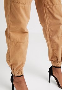 Pantalons cargo kaki fuselés avec des poignets élastiques aux chevilles, fabriqués dans un tissu doux et durable. Accompagnés d'une chaussure noire à talons hauts.