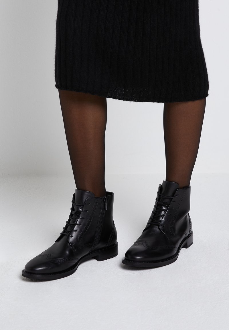 Botas de tobillo de cuero negro con cordones en la parte delantera y cremallera lateral. Presentan detalles de brogue en la puntera y un tacón corto apilado.