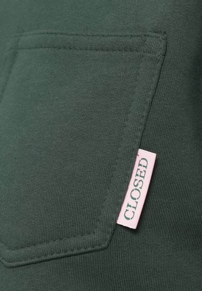 Το ύφασμα της πράσινης μπλούζας φούτερ διαθέτει μια λοξή τσέπη με ροζ ετικέτα που γράφει "CLOSED". Η υφή είναι λεία με ελαφριά ελαστικότητα.
